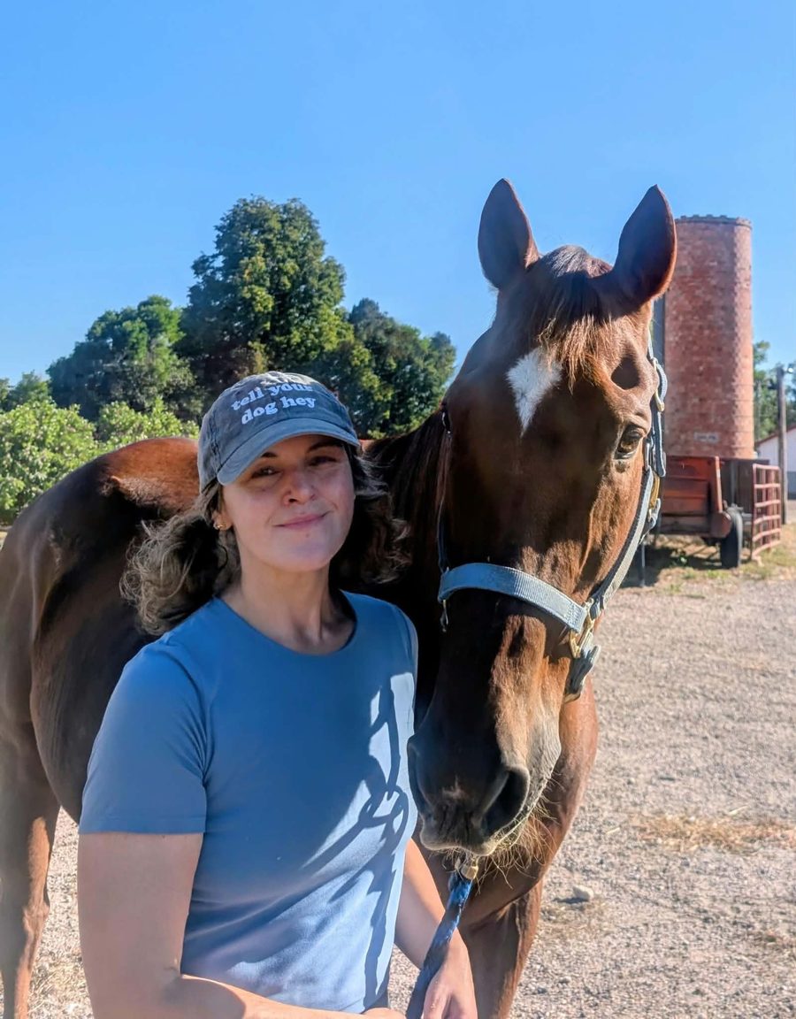 Woman in a ball cap standing beside a chestnut horse with a white star, on a sunny farm
