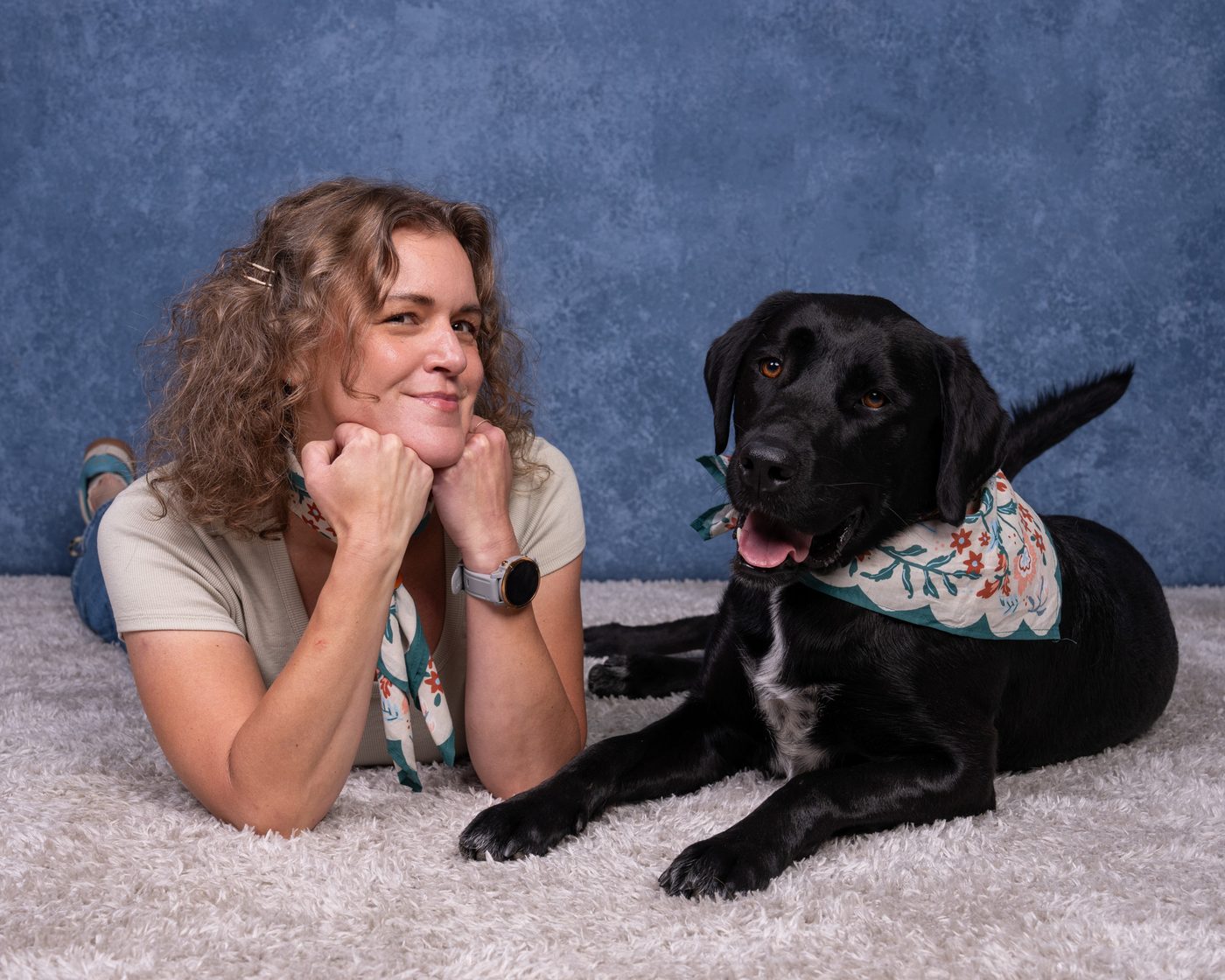 Woman lying on a rug beside a black therapy dog, both wearing matching floral bandanas