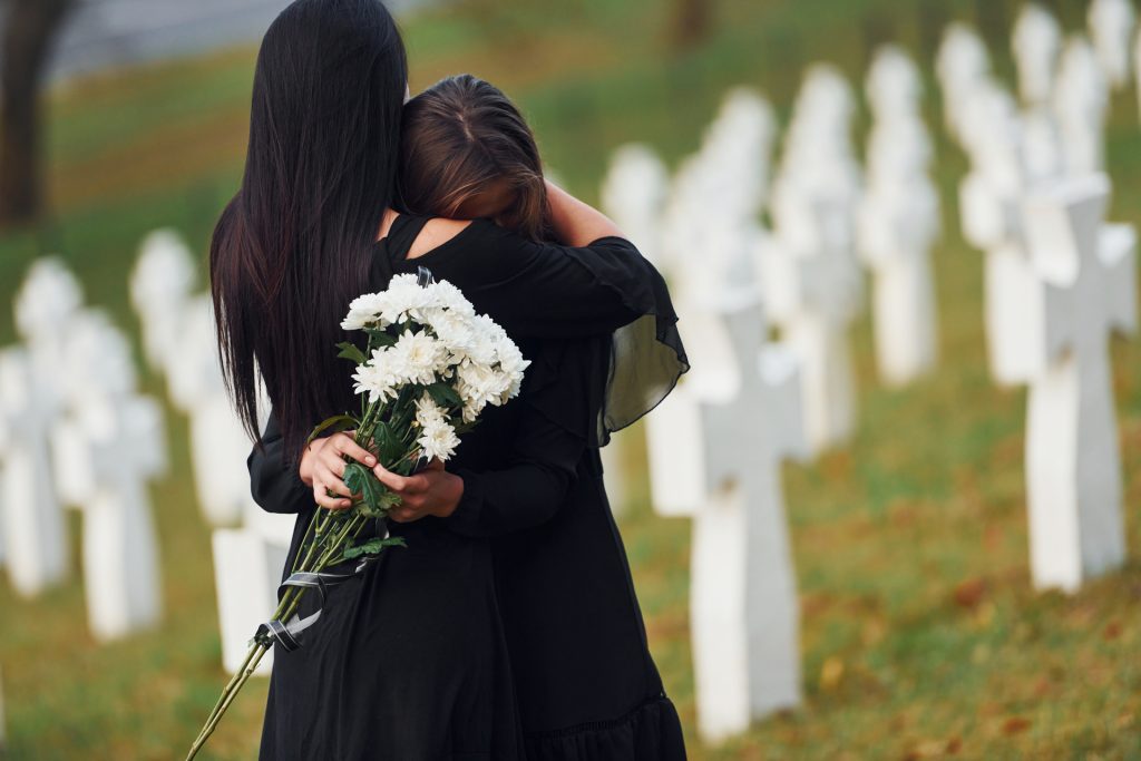 embracing each other and crying. two young women in black clothes visiting cemetery with many white crosses. conception of funeral and death