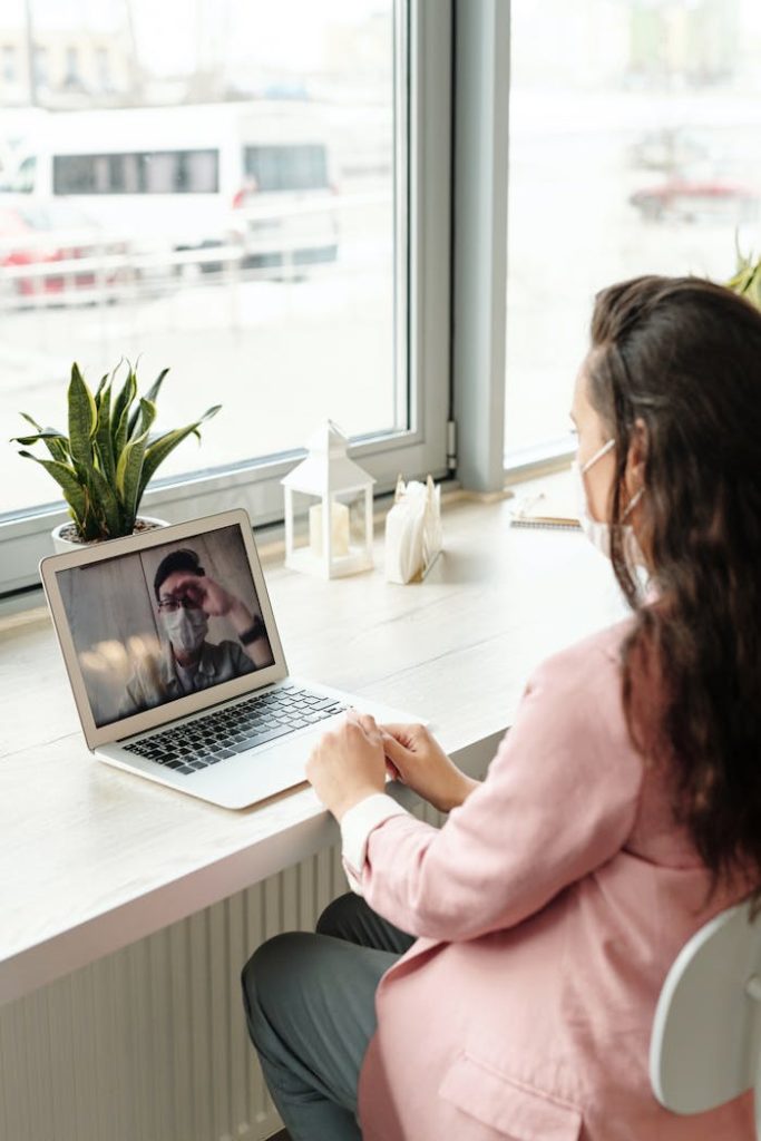 A woman wearing a face mask participates in a video call from her home office, showcasing remote work during the pandemic.